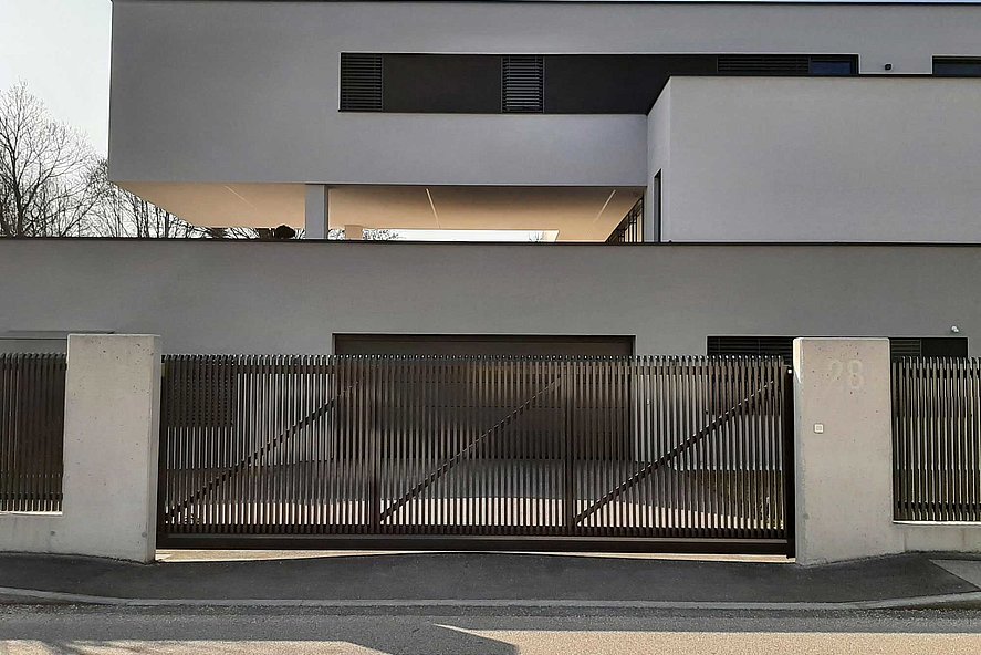 Schiebetor im Stil eines Lattenzauns mit Schrägkappen vor einer Einfahrt eines modernen Einfamilienhauses