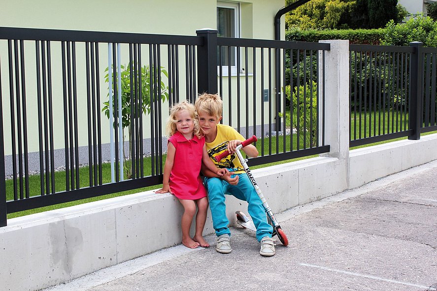 Gartenzaun aus Alumium mit 3-fach Stabreihen in anthrazit auf weißem Mauersockel, davor zwei Kinder Wange an Wange mit Roller