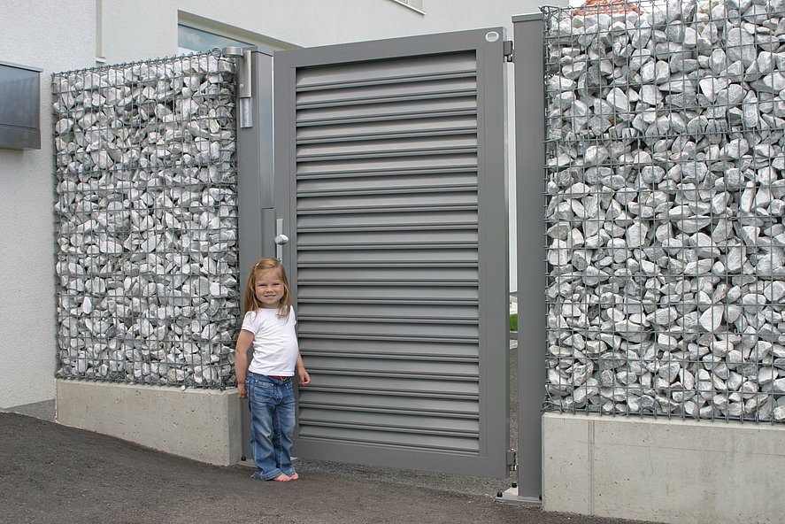Gartentür mit Lamellen in grau als Eingang zum Gabionenzaun mit kleinem Mädchen in der Einfahrt
