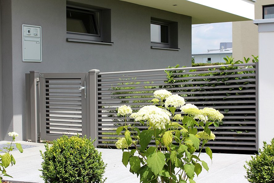 Gartentür mit Zaunelement eines Lamellenzauns aus Aluminium in edlem grau mit Büschen davor