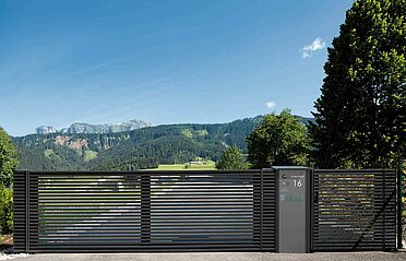 Modernes Schiebetor aus Aluminium und dazu passender Briefkastenäule begrenzt eine lange Einfahrt, im Hintergrund ist ein Einfamilienhaus zu sehen