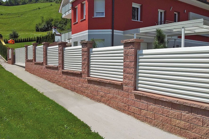 weißer Gartenzaun mit Lamellen auf Mauersockkel aus Backsteinen umzäunt ein Appartmenthaus in den Bergen