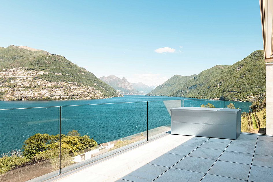 Moderne Gartenbox aus Stahl in silber metallic steht auf einem Balkon mit Aussicht auf einen See