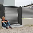 Junge lehnt an Gartentür mit grauen Lamellen aus Aluminium auf weißem Mauersockel